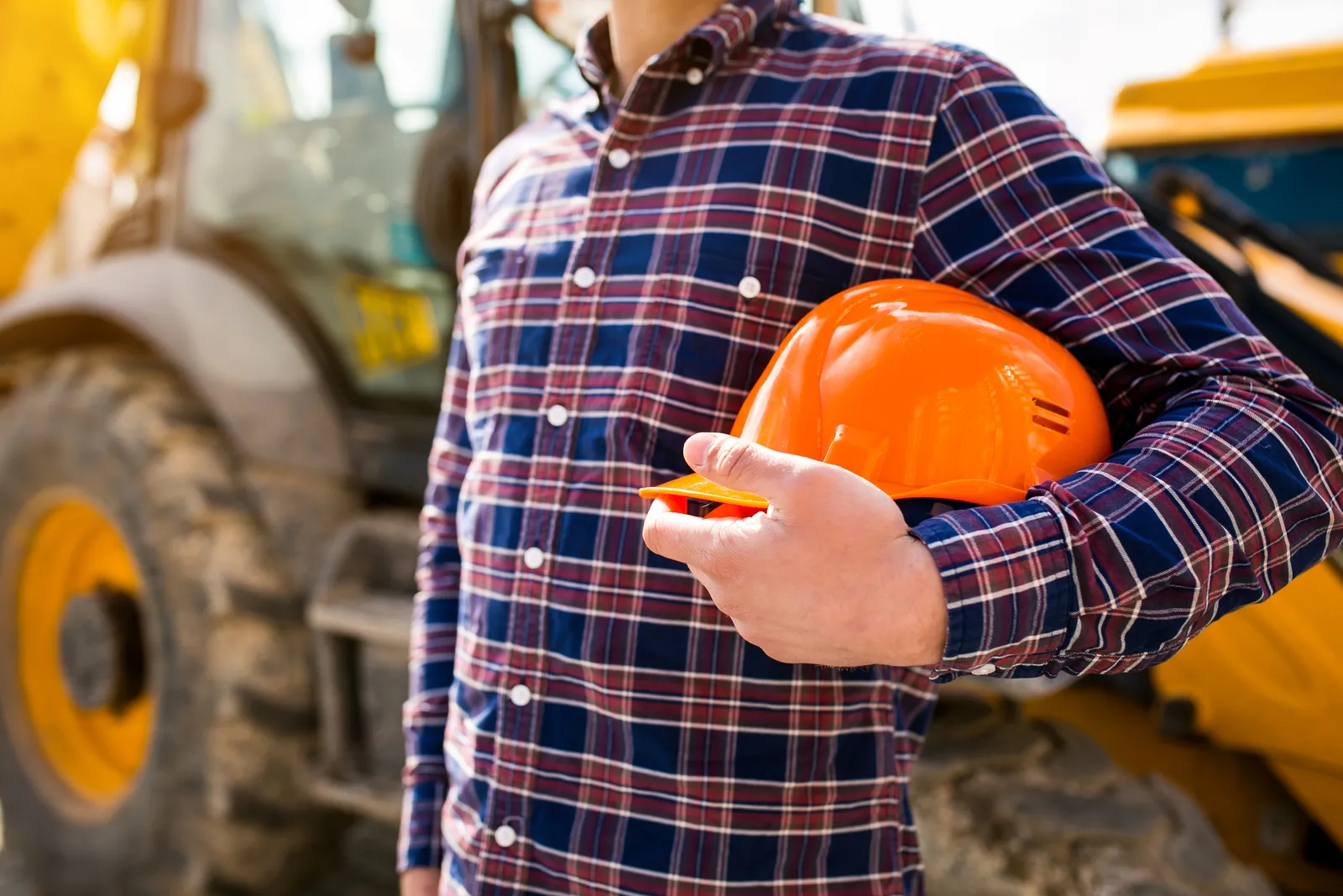 Operatore del settore edile con casco di protezione arancione davanti a una macchina movimento terra in cantiere.