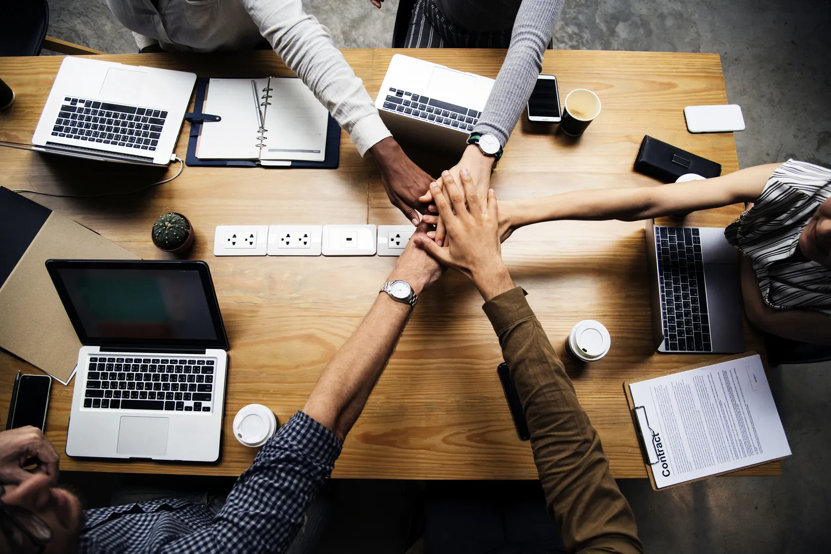 Gruppo di persone che unisce le mani durante una riunione di lavoro attorno a un tavolo con computer e strumenti digitali